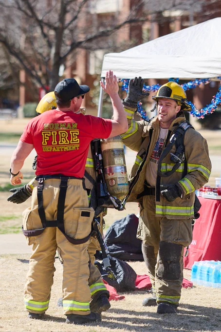 Two uniformed firefighters, one wearing a "Norman Fire Department" shirt, exchange a high-five under a canopy. The scene is outdoors, with firefighting equipment visible. A refreshment table, decorated with blue garland, is set up nearby.