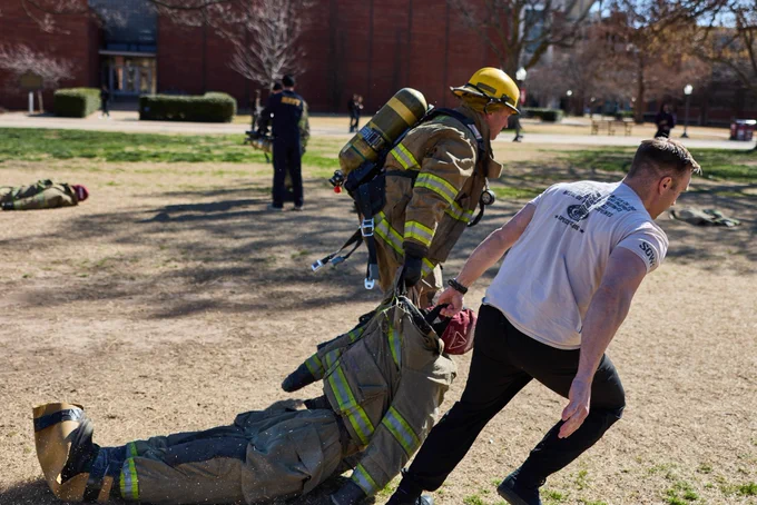 A person in a firefighter suit and helmet, carrying equipment, drags a firefighter dummy across a grassy area. Another person in a T-shirt assists with the relay game. Additional people and buildings are visible in the background.