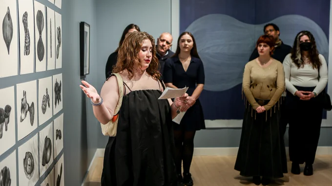 A female student speaks in front of a gallery wall of lithograph prints at the the "speculative morphologies" exhibition opening.