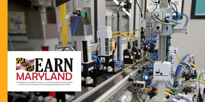 Automated industrial machinery with robotic arms and conveyor belts in a manufacturing setting. EARN Maryland logo in bottom left corner.