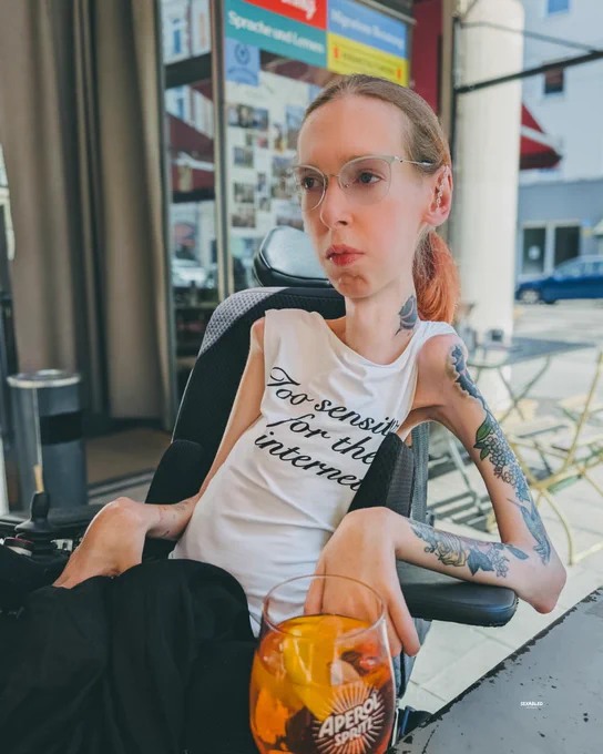Das Foto zeigt mich an einem grauen Tisch im Außenbereich eines Cafés. Ich trage ein weißes Tanktop mit der Aufschrift „Too sensitive for the internet“, eine schmale Brille und meine rötlichen Haare zurückgebunden. Meine Tätowierungen an Hals und Armen sind deutlich sichtbar. Im Vordergrund steht ein Glas Aperol Spritz. Ich blicke mit einem ernsten, entschlossenen Ausdruck leicht an der fotografierenden Person vorbei. Der Hintergrund zeigt eine Stadtstraße mit einem Gebäude der „Asylberatung“.
