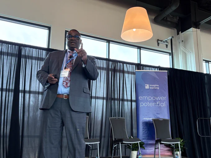 A man is delivering a Keynote address on stage. And imagine learning banner is behind him. The banner reads and empower potential.