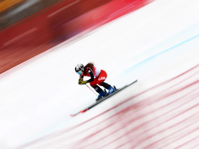 Mollie Jepsen in a red and black outfit, with "CAN" visible on the suit, speeds down a snowy slope. The background is motion-blurred, emphasizing the sense of speed.
