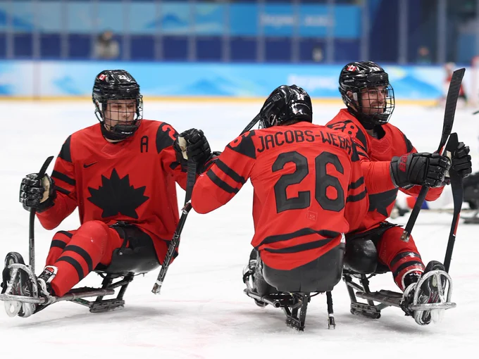 Three athletes in red and black jerseys, featuring the Maple Leaf, are playing Para ice hockey on ice. One player's jersey displays the name "Jacobs-Webb." They are actively engaged in gameplay, using their sticks to propel themselves. The scene takes place in an arena with a blue-themed backdrop.