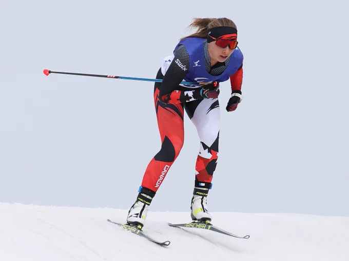 Natalie Wilkie, a Cross-country skier in dynamic motion on a snowy slope, wearing a suit with the word "CANADA" and using ski poles. The skier is focused, with a determined posture, and is dressed in a mix of red, black, and white, with a blue bib.