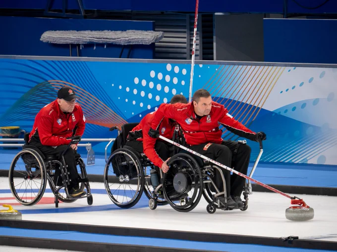 Three athletes in wheelchairs are playing a game of wheelchair curling on ice, wearing red uniforms. Mark Ideson is pushing a curling stone with a stick while the others watch. The background features a blue and white abstract design.