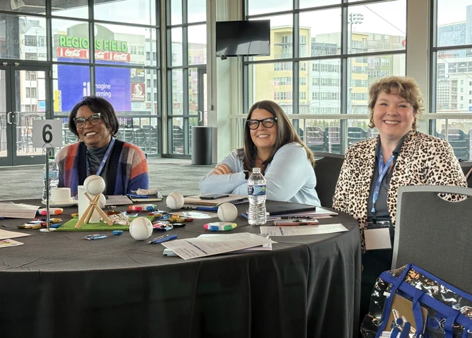 Three women are smiling at the camera. They are sitting behind a table during a conference. In the background, you can see the regions field sign.
