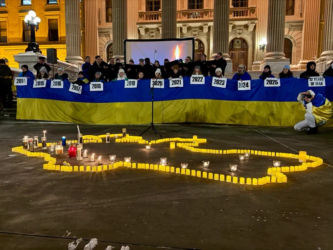 A group of people stand in front of a Ukrainian flag, holding signs with years from 2014 to 2025. They are gathered outside the Alberta Legislature Building, and a small stage has a screen displaying a candle. In front, a map outline of Ukraine is made with lit candles.