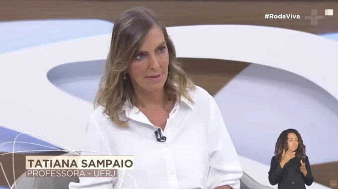 A imagem é uma captura de tela da participação da professora Tatiana Sampaio no programa de tevê “Roda Viva", da TV Cultura.  A imagem mostra a professora no estúdio de televisão, enquadrada do peito para cima, durante a entrevista. Ela veste uma camisa branca de mangas longas, com um microfone de lapela preso à roupa. Ela é branca e possui cabelos claros, de comprimento médio, levemente ondulados. Sua expressão facial é atenta e séria, com o olhar direcionado para alguém fora do enquadramento, sugerindo que está ouvindo ou respondendo a uma pergunta. Na parte inferior da tela, há um gerador de caracteres com o nome Tatiana Sampaio e a identificação Professora – UFRJ.