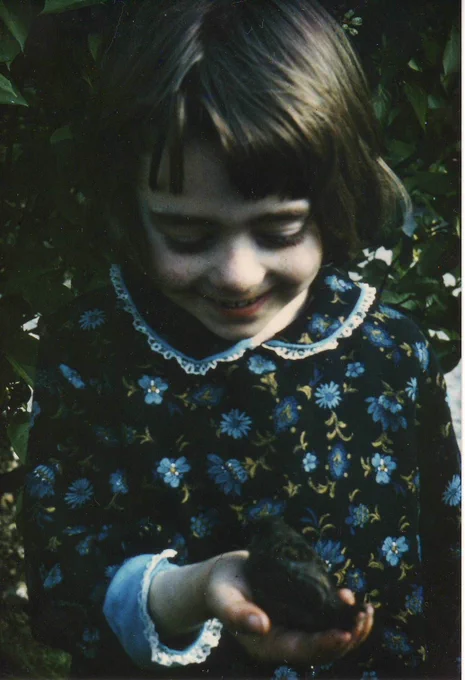 A beautiful timeless colour photograph of young Penny aged about five years old. Shown from the waist up, Penny holds a small dark bird in one hand, as it nestles on her palm. She is wearing a handmade dress, dark blue with pale blue cornflowers and a  cute lace trimmed collar. Her brown hair is cut in a bob with an unruly fringe. She's looking down and smiling at the bird. In the background there is dark green foliage.