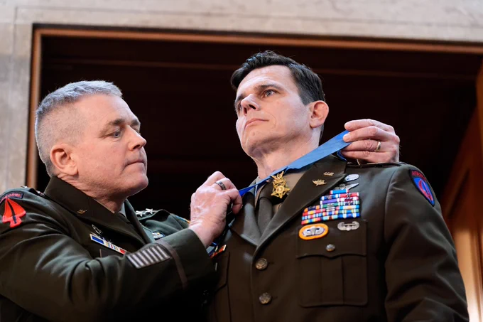 A military officer awards a soldier a medal, draping it around his neck. Both wear decorated uniforms, conveying honor and solemnity in a formal setting.