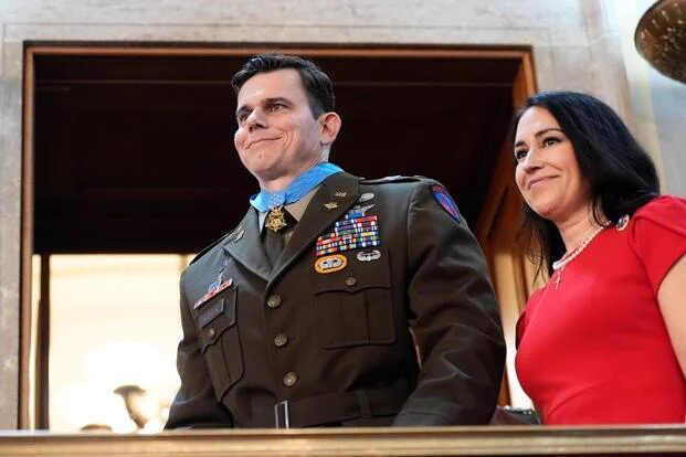 A person in a military uniform with various medals and badges stands next to another person wearing a red outfit. They are in a formal setting, possibly a government or official building.