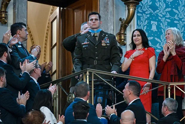 A person in a military uniform, wearing numerous medals, stands in a balcony area surrounded by applauding individuals. Nearby, another person in a red outfit places a hand on their chest. The setting appears formal, with ornate architectural details and a patterned blue wall.