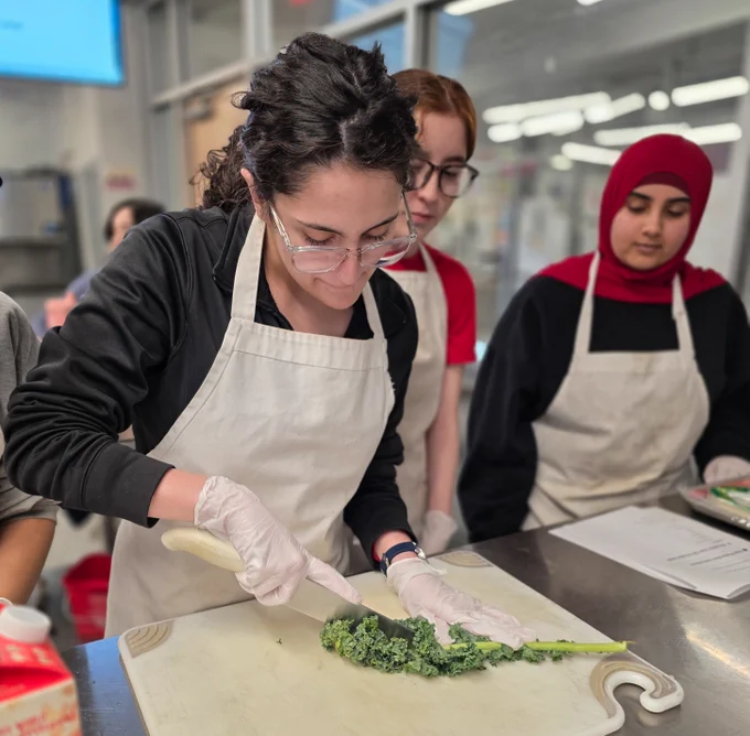 A JTED Culinary Arts student watches two Medical Assistant students cut leafy greens to prepare fresh and nutritious food
