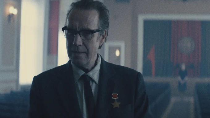 From Star City, Rhys Ifans as Chief Designer looks ahead while wearing a suit and glasses.
