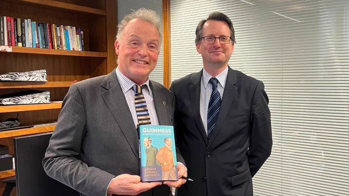 Arthur Edward Guinness, 4th Earl of Iveagh, stands on the left holding a copy of his book Guinness: A Family Succession, beside Professor Patrick Geoghegan, Director of the Trinity Long Room Hub, on the right. They are pictured indoors in front of wooden bookshelves and office blinds.
