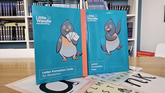 Image of Little Wandle Handwriting Letter Formation Cards for Reception and Letter Formation Mats standing up on top of a flat lay of cards and mats. The background shows a brown table and a book case.