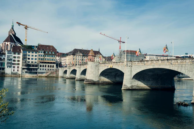 A picturesque scene of Basel's historic bridge with city buildings and cranes by the Rhine River.