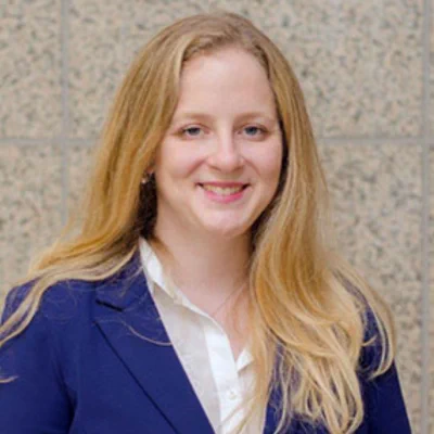 A woman with long blonde hair smiling at the camera, wearing a navy blazer over a white blouse, standing in front of a light stone wall background.