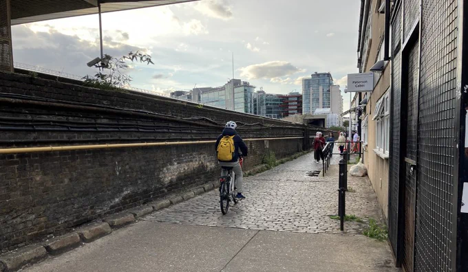 Cyclist wearing a yellow backpack rides along a cobblestone path beside a brick wall under a cloudy sky in an urban area.