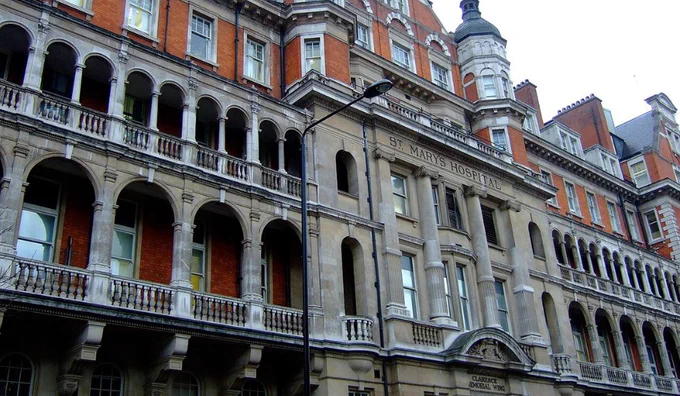 Facade of St. Mary's Hospital featuring ornate stone balconies and red brickwork under a cloudy sky.