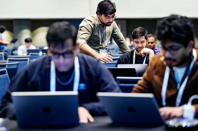 Two people working on laptops at a table, and one person consulting another working on their laptop in the background at KubeCon   CloudNativeCon