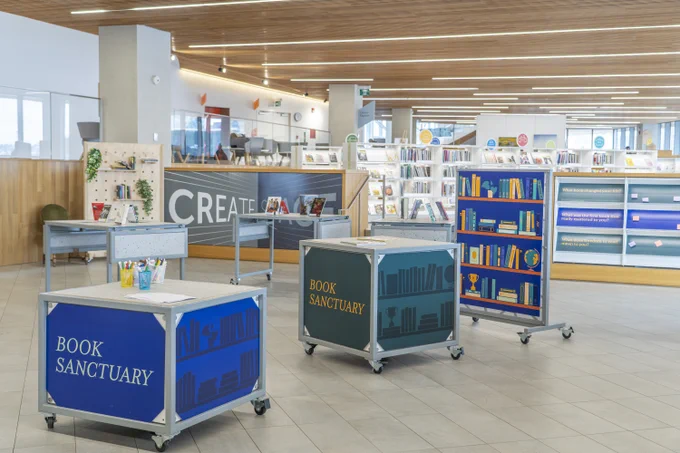 A library interior with wooden ceilings and large windows. In the foreground, there are mobile carts labeled "Book Sanctuary." In the background, there are more shelves with books and information on posters. 