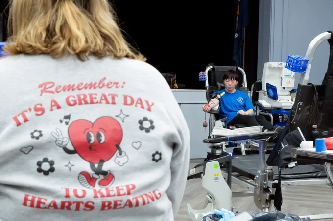 The back of a shirt that reads "Remember: It's a great day to keep hearts beating," and a student in the background donating blood during the blood drive on campus. 