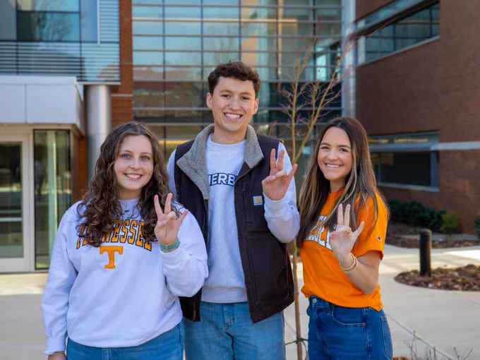 Three students stand smiling outside a university building, wearing University of Tennessee apparel and making the "Vols" sign with their hands.