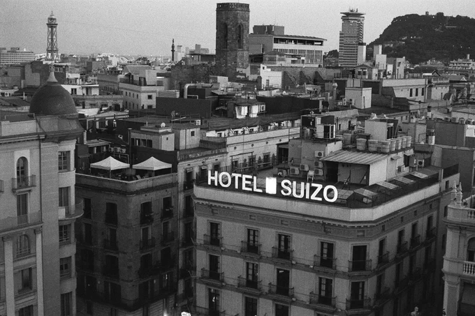 A black and white cityscape featuring the Hotel Suizo sign prominently in the foreground. The view includes numerous surrounding buildings and rooftops, with notable architectural elements. In the background, a hill with a structure atop it can be seen.