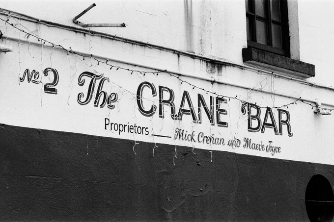 Black and white photo of painted sign for "No 2, The CRANE BAR," with decorative string lights and smaller text below reading, "Proprietors Mick Crehan and Maeve Joyce."