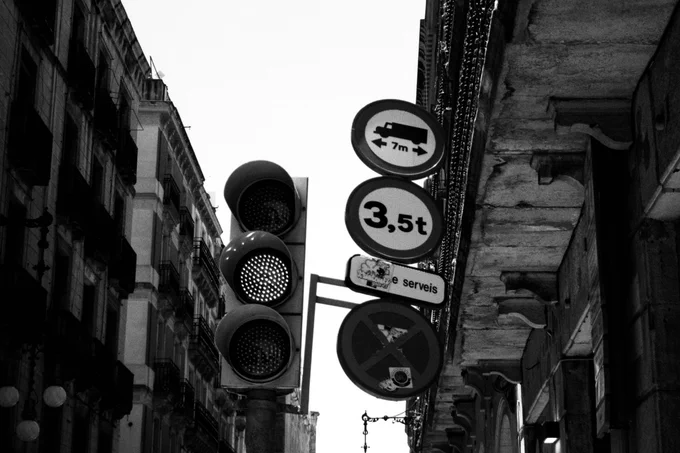 A street view shows a traffic light and several traffic signs mounted on a pole, including weight and height restrictions. Buildings and balconies are visible on both sides of the street.