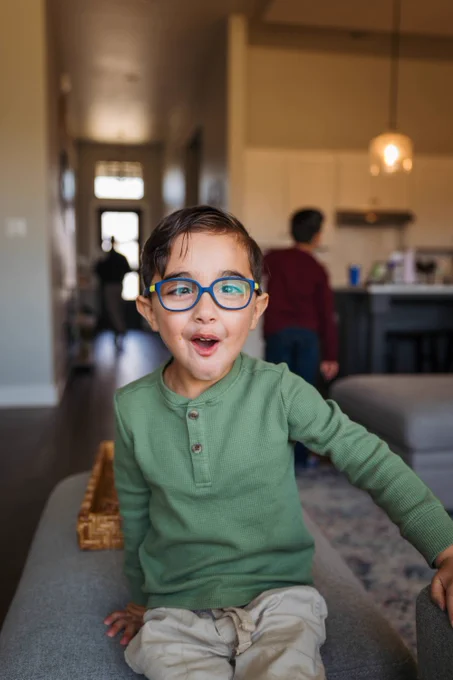 A child with short dark hair and blue glasses is sitting on a sofa, looking excitedly at the camera. The child is wearing a green long-sleeve shirt and light-colored pants. In the background, another child stands near a kitchen area. The setting appears to be a modern home with an open floor plan.