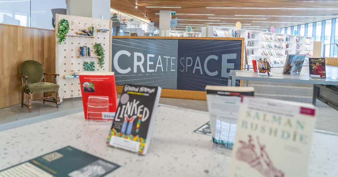 A library interior featuring a "Create Space" sign on a partition. Various books are displayed in the foreground. The background shows shelves filled with books and a small reading area with a green chair and display of books and plants.
