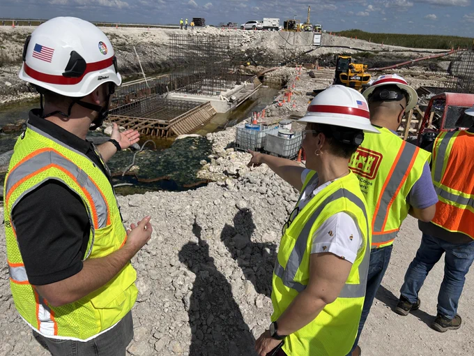 Miguel Lugo and a colleague from the US Army Corps of Engineers are in bright safety vests and hard hats are conversing at a construction site. They are near a partially built structure with rebar and concrete forms. Other colleagues and constructions workers and construction equipment are visible in the background. An American flag is on one helmet. The setting appears to be a large infrastructure project.