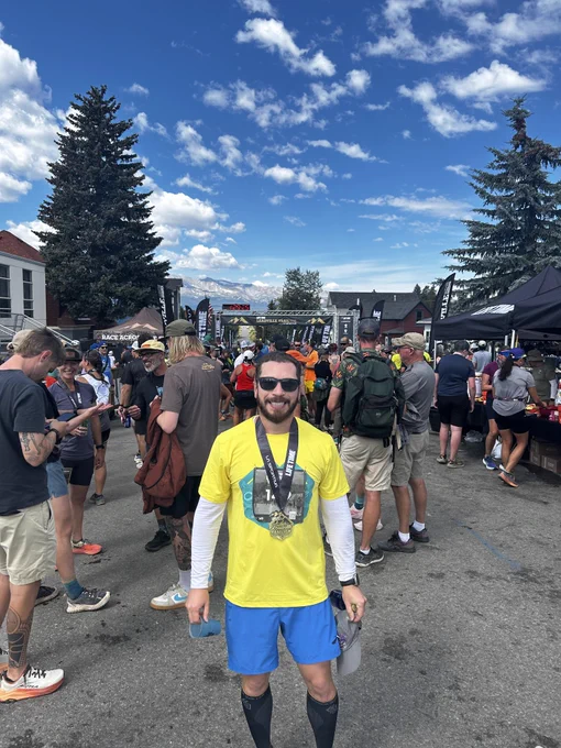 Miguel Lugo is in a bright yellow shirt and blue shorts stands proudly in the foreground, wearing a medal. Behind him, a crowd gathers at an outdoor event with various tents and banners. The sky is blue with clouds, and the setting includes trees and buildings.