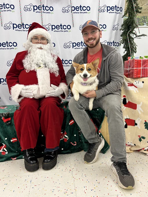 Miguel Lugo who is holding his dog, Pepa, is sitting next to a person in a Santa suit who is seated on a festive bench. They are in front of a backdrop with the Petco logo and surrounded by holiday decorations, including wrapped gifts and a blanket with Christmas motifs.