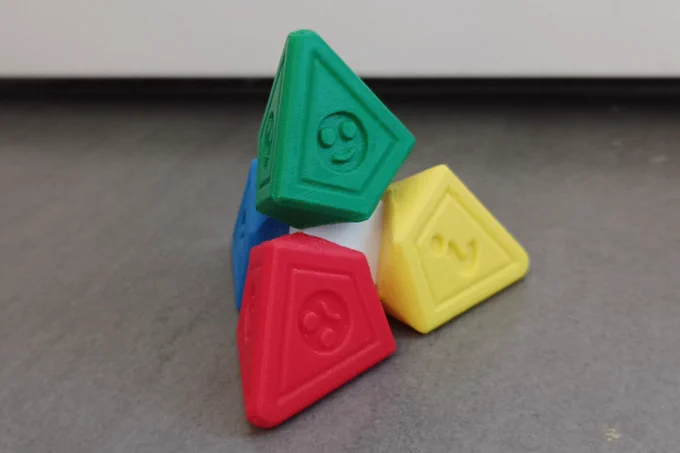 Photo prise par les élèves du collège Jacques Prévert de Noisy-le-Sec. La photo est composée d'un fond gris et d'un objet imprimé en 3D en forme de tétraède. Chaque extrémité du tétraède est d'une couleur différente (verte, rouge, jaune et bleue), avec des pictogrammes de visages souriants ou non gravés sur chaque face. Cet objet est utilisé en classe pour signaler son état ou son besoin. 