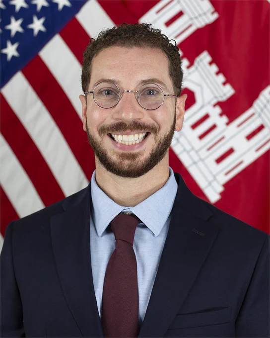 Miguel Lugo has a suit on and is smiling, standing in front of an American flag and a US Corps of Army Engineers flag.