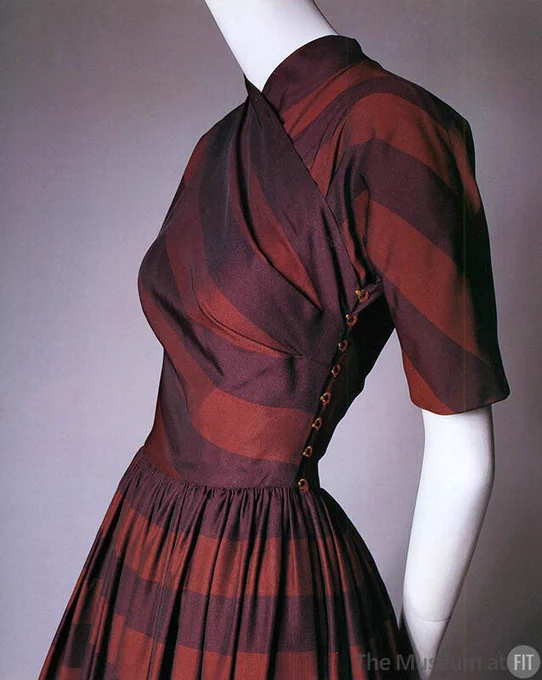 A view of the bodice of a purple and red striped gown with a high crossover treatment at the neck, sleeves above the elbow and a line of ball style buttons that fasten at the side of the waist