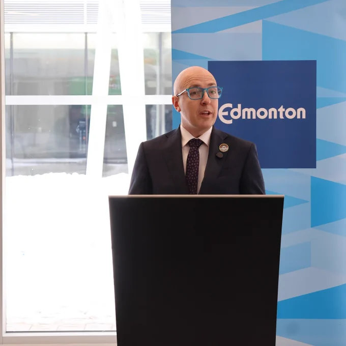 Mayor Knack is speaking at a podium indoors. Behind the podium is a background with the word "Edmonton" on it.