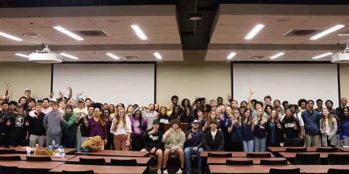 A large group of people posing together in a classroom setting. They are standing in front of empty desks and waving at the camera.