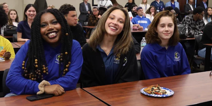 A group of SPC students is seated in a classroom setting with multiple tables. Three people in the foreground are smiling at the camera. Some people in the background are engaged in conversation. 