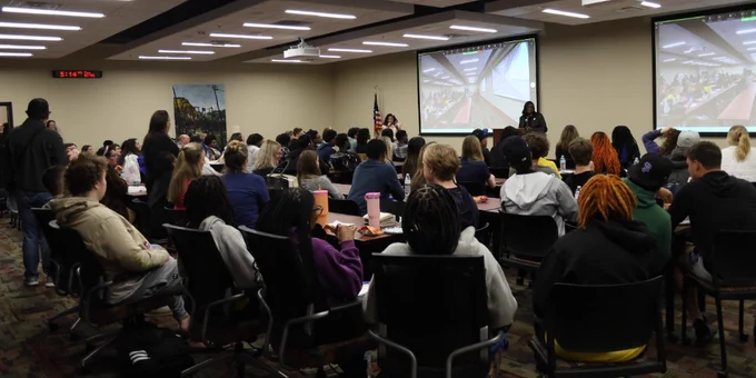 A large group of SPC students is seated in a conference room facing two projection screens. One person is standing near the front, speaking to the audience.