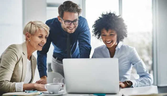 A diverse team of professionals collaborating and smiling while working on a laptop in a modern office setting.