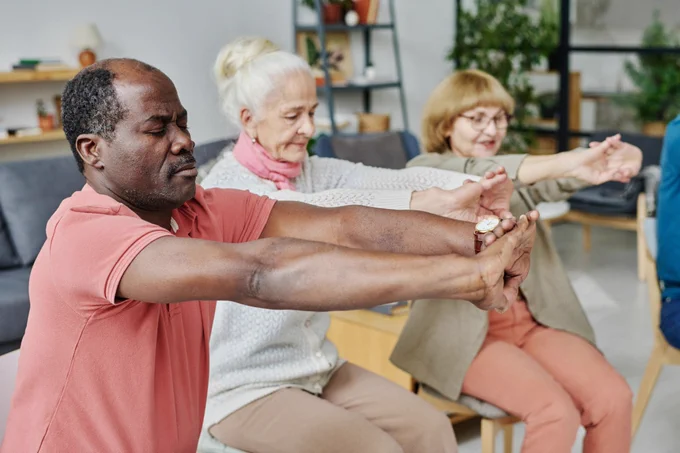 A group of senior citizens sit in chairs in a circle and stretch their arms out in front of them. 