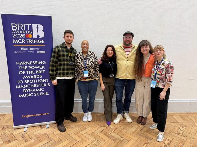 A group of six people standing indoors in front of a "Brit Awards 2026 MCR Fringe" banner. They are smiling and appear to be at an event. The floor is patterned wood, and the banner has text promoting the Brit Awards in Manchester.