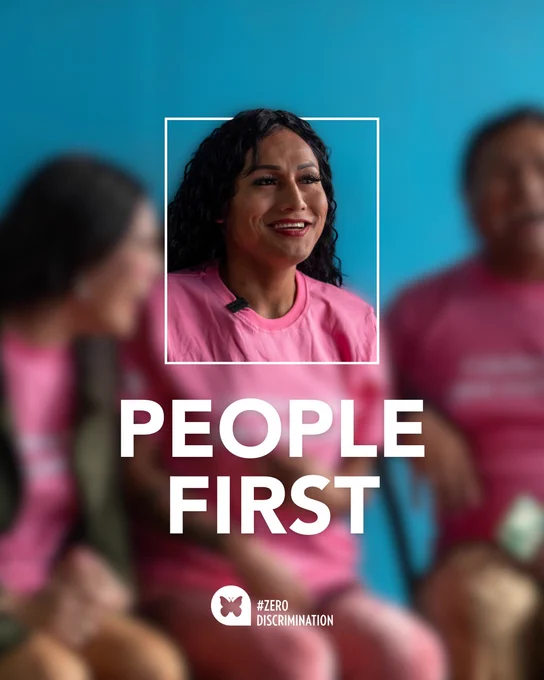 Image of 3 people seating next to each other. The face in the middle is in focus while the surrounding people and background are softly blurred, drawing attention to the central subject. A white rectangular frame surrounds the person, and bold white text below reads “PEOPLE FIRST,” accompanied by the #ZeroDiscrimination logo
