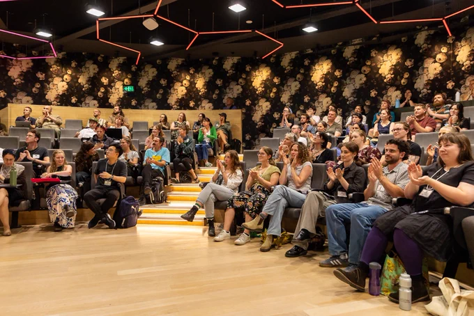 A photo of a full lecture theatre with light wooden floors, a dark back wall and red LED lights in the shape of a honeycomb above the audience.