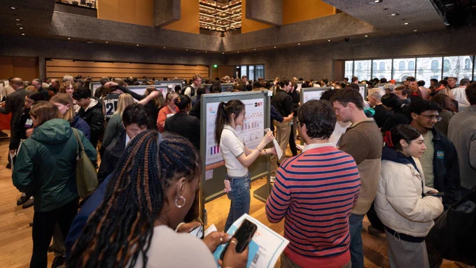 A busy conference hall filled with people viewing and discussing research posters. The atmosphere is lively, with attendees engaged in conversation.